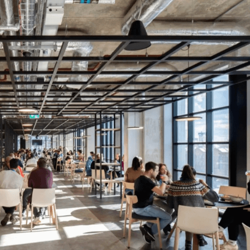 Modern office cafeteria with exposed HVAC ductwork and large floor-to-ceiling windows.